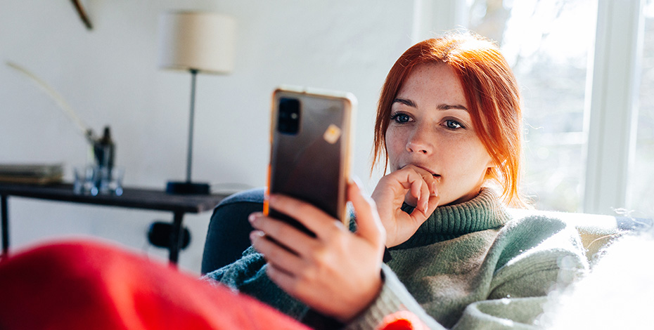 A troubled woman lay on the couch with a blanket over her while reading something intently on her mobile phone.