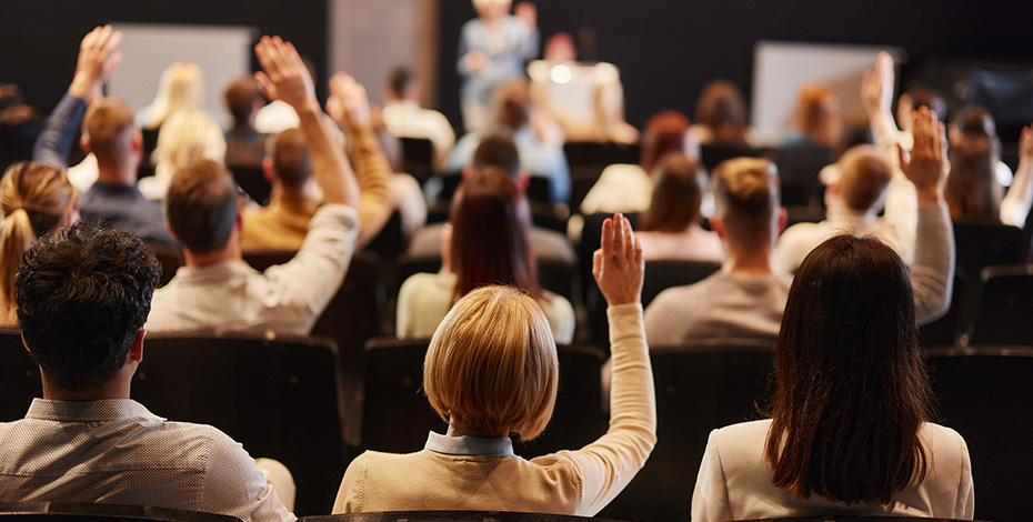 A view of people at a conference from behind them. Some are raising their hands. 