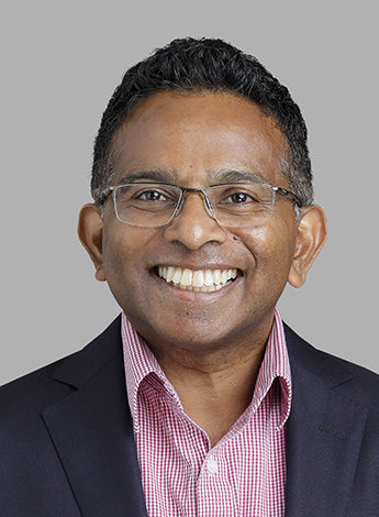 A dark-skinned man with short dark brown hair and glasses is looking at the camera