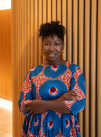 "A woman of African descent wearing brightly coloured clothes is standing in front of a timber wall with her arms crossed. "