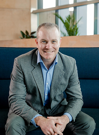 "A man wearing a grey suit is sitting on a blue couch. There is a window and some plants behind him."