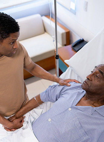 Young man comforts older man in hospital bed.